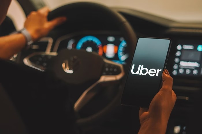 Motorista de aplicativo Uber segurando smartphone com logo da empresa no painel do carro, representando o lucro recorde de R$ 9 bilhões em 2025